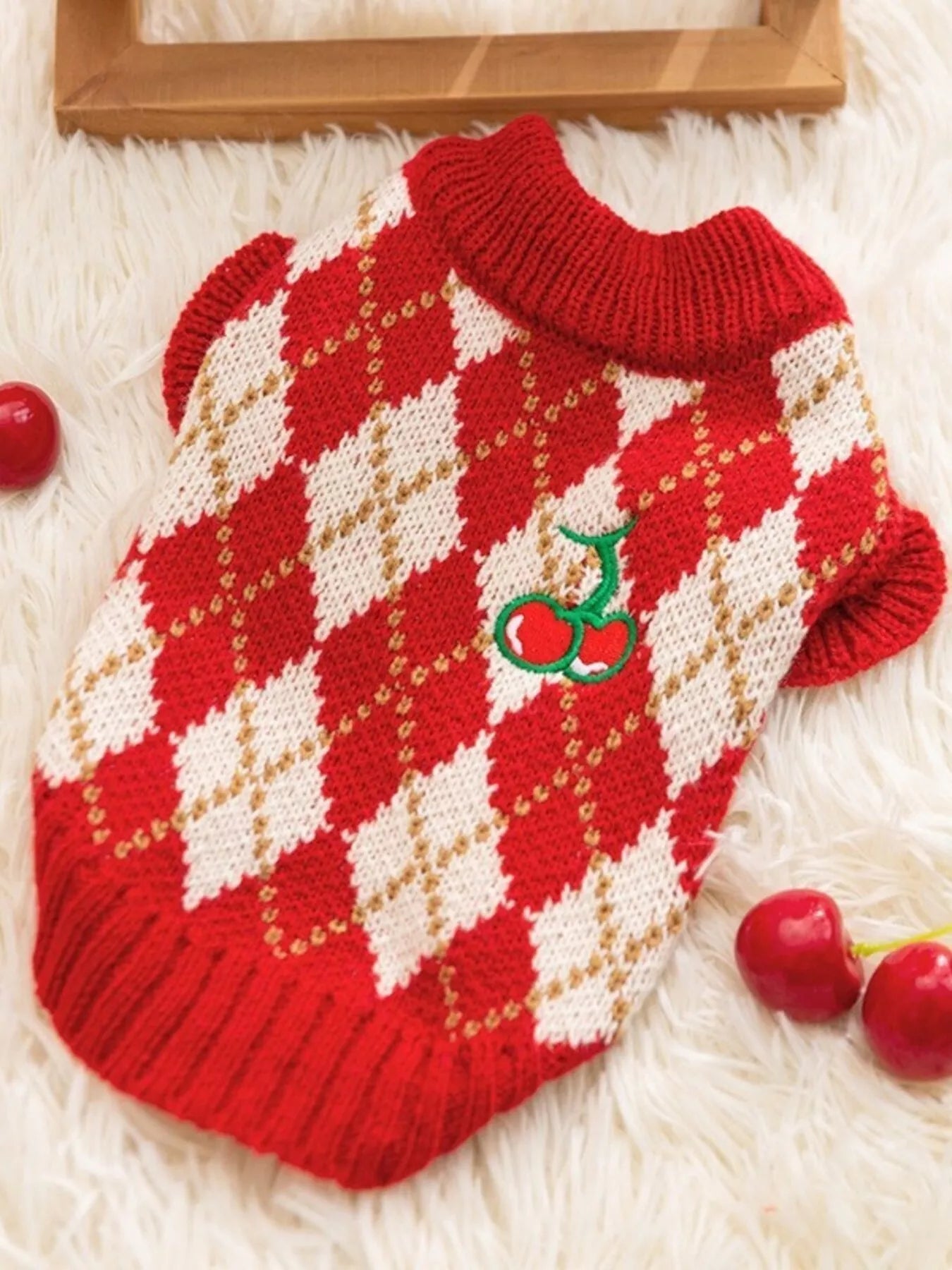 Red and white argyle patterned dog sweater with cherry design on a fluffy white surface.