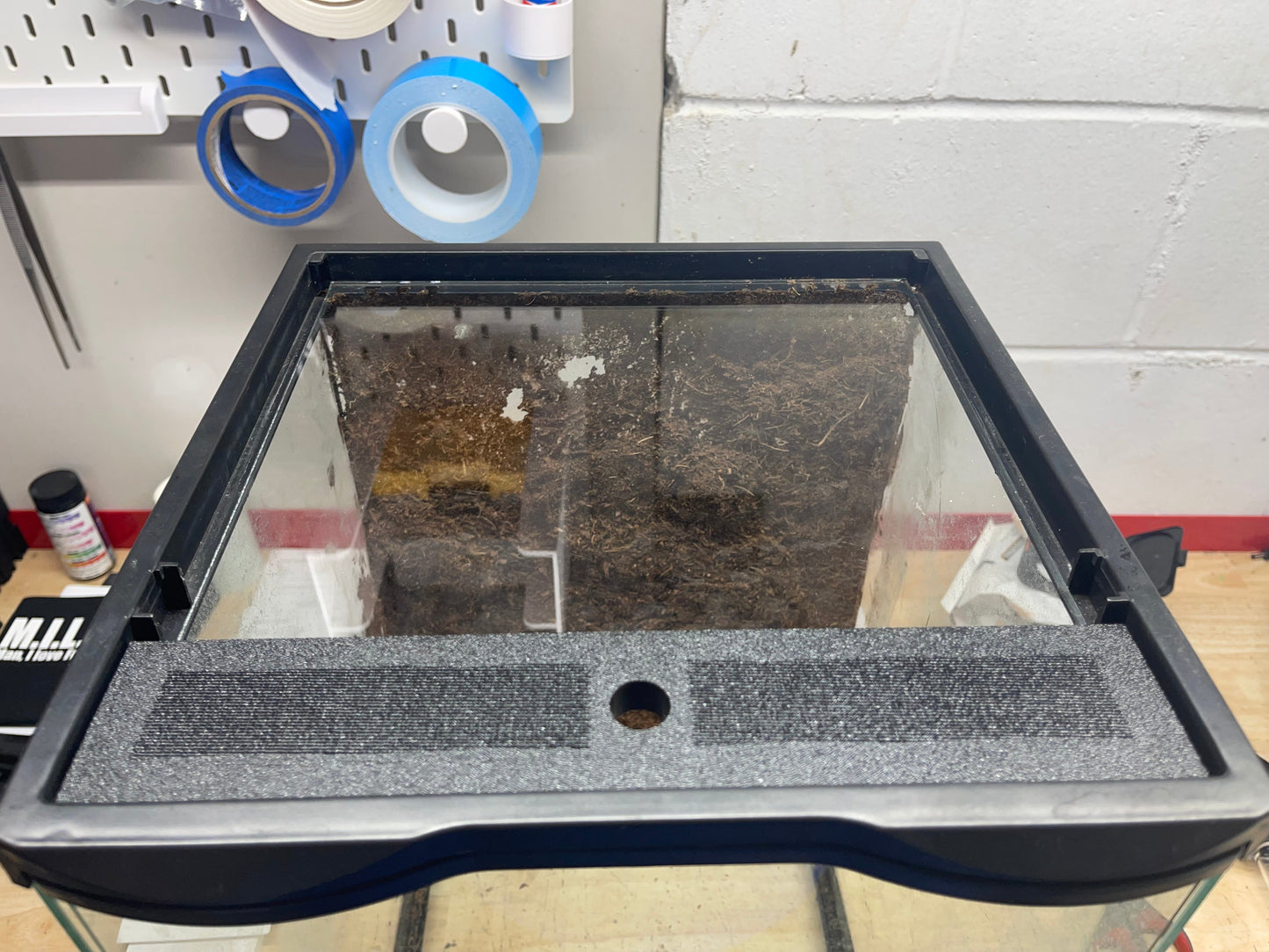Glass display case with a black frame on a wooden floor, with tape and a container in the background.