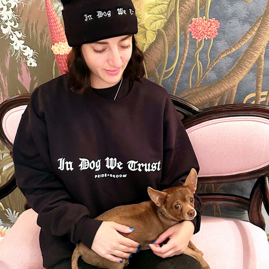 Person wearing a black 'In Dog We Trust' sweatshirt holding a small dog, sitting on a decorative chair with floral and abstract patterns.