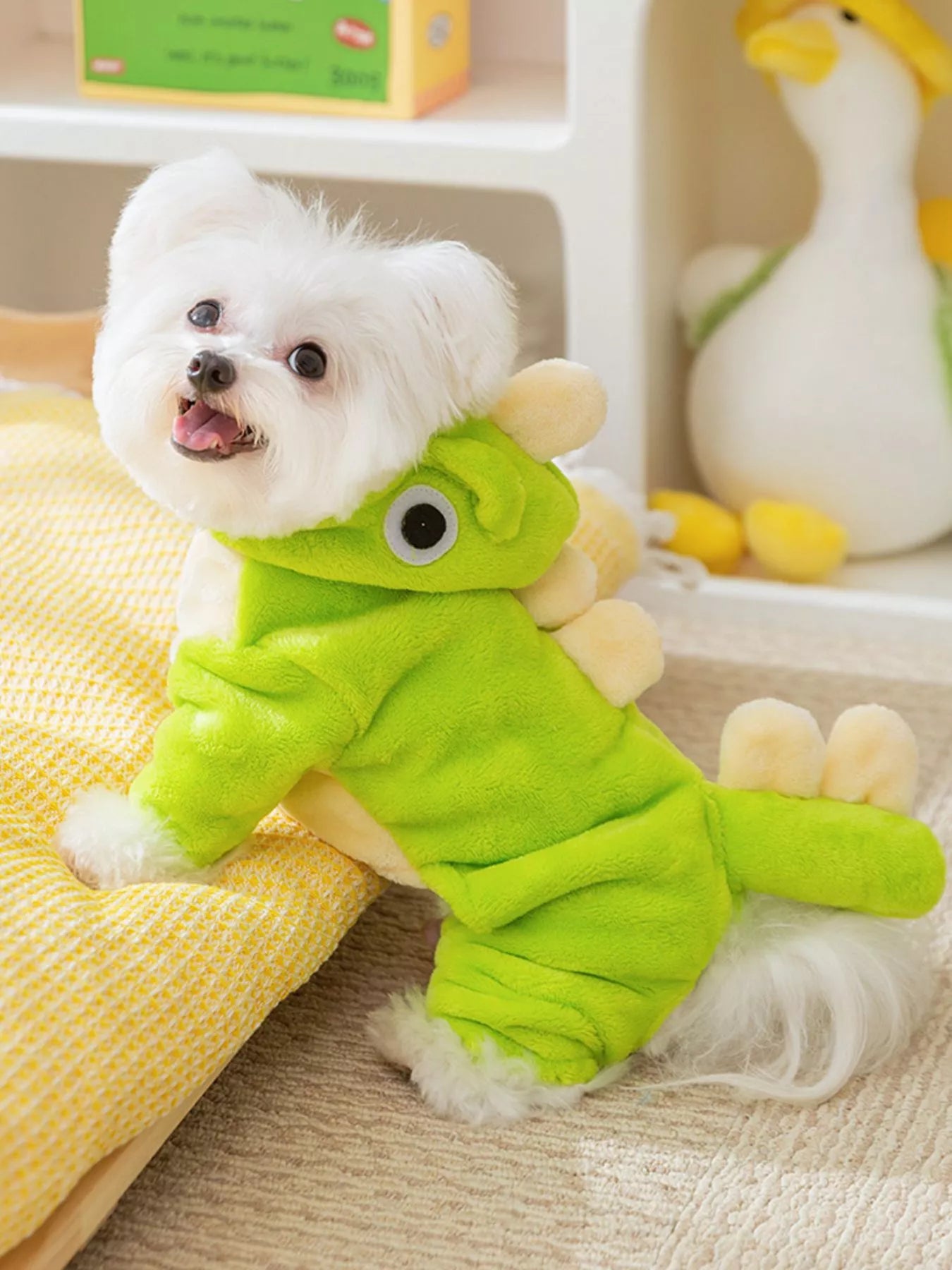 Small white dog wearing a green dinosaur costume on a wooden floor.