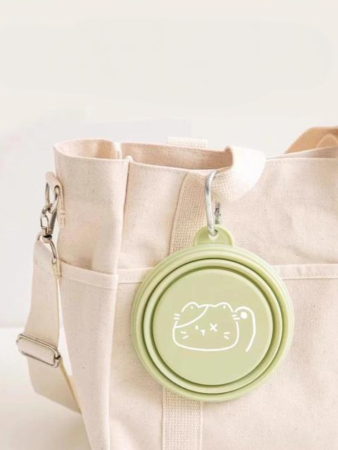 Green collapsible cup with a cat design attached to a beige tote bag on a light background