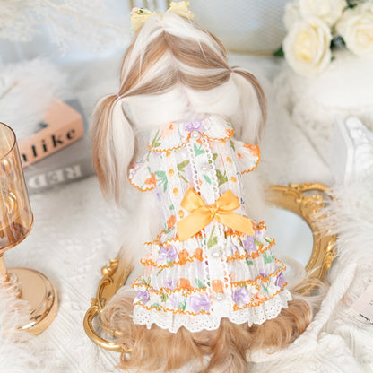 Small doll wearing a floral dress with a yellow bow, sitting on a decorative surface.