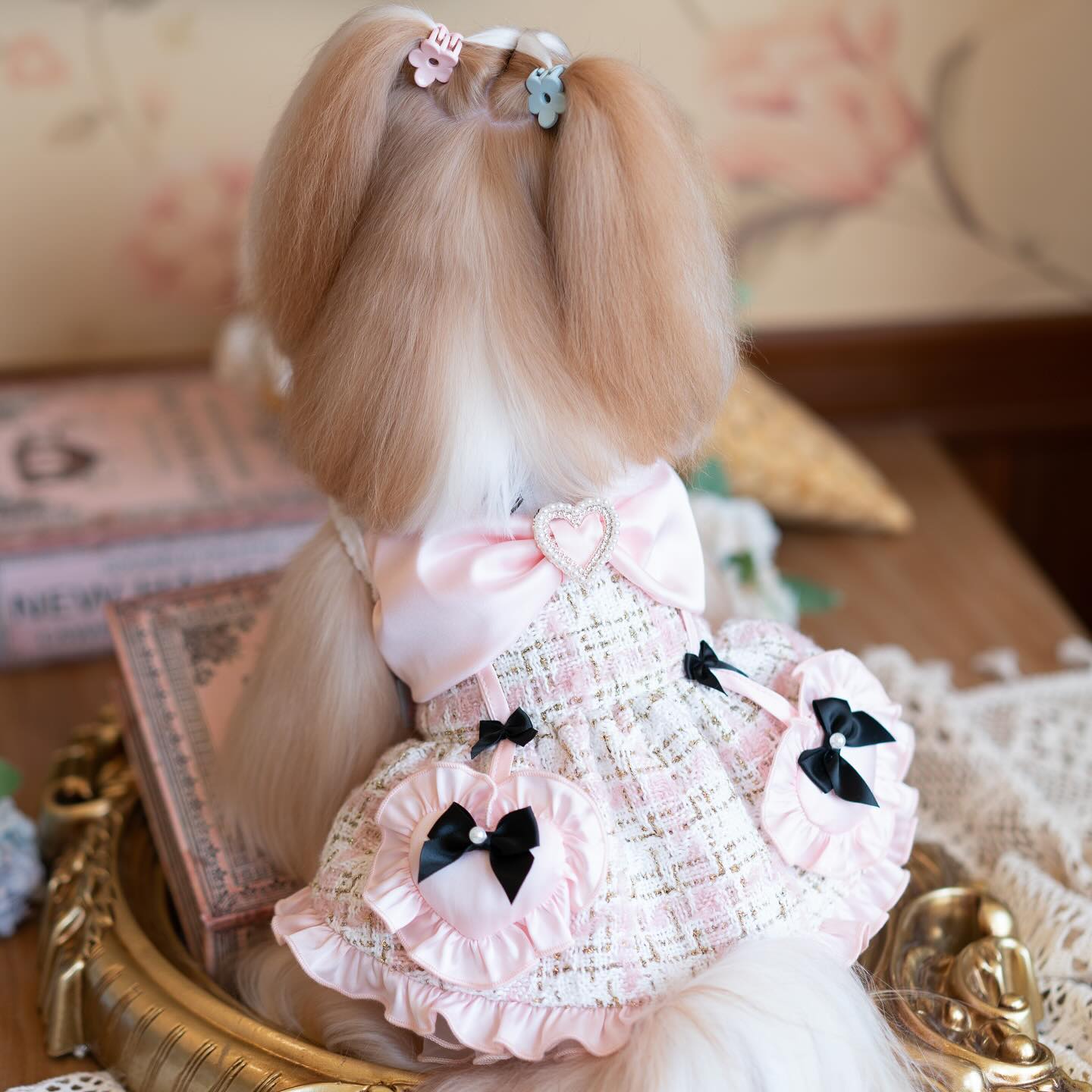 Doll with blonde hair styled in pigtails, wearing a pink dress with black bows, in a decorative setting.