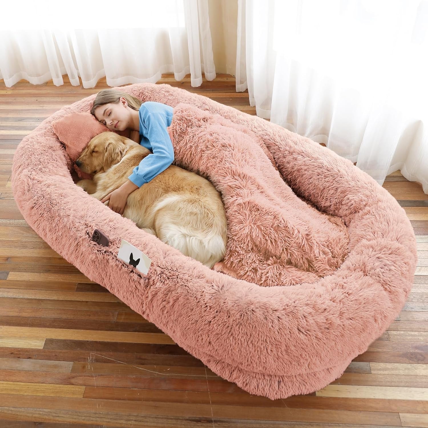 Woman hugging a large pink fluffy dog bed with a golden retriever inside, in a room with curtains.