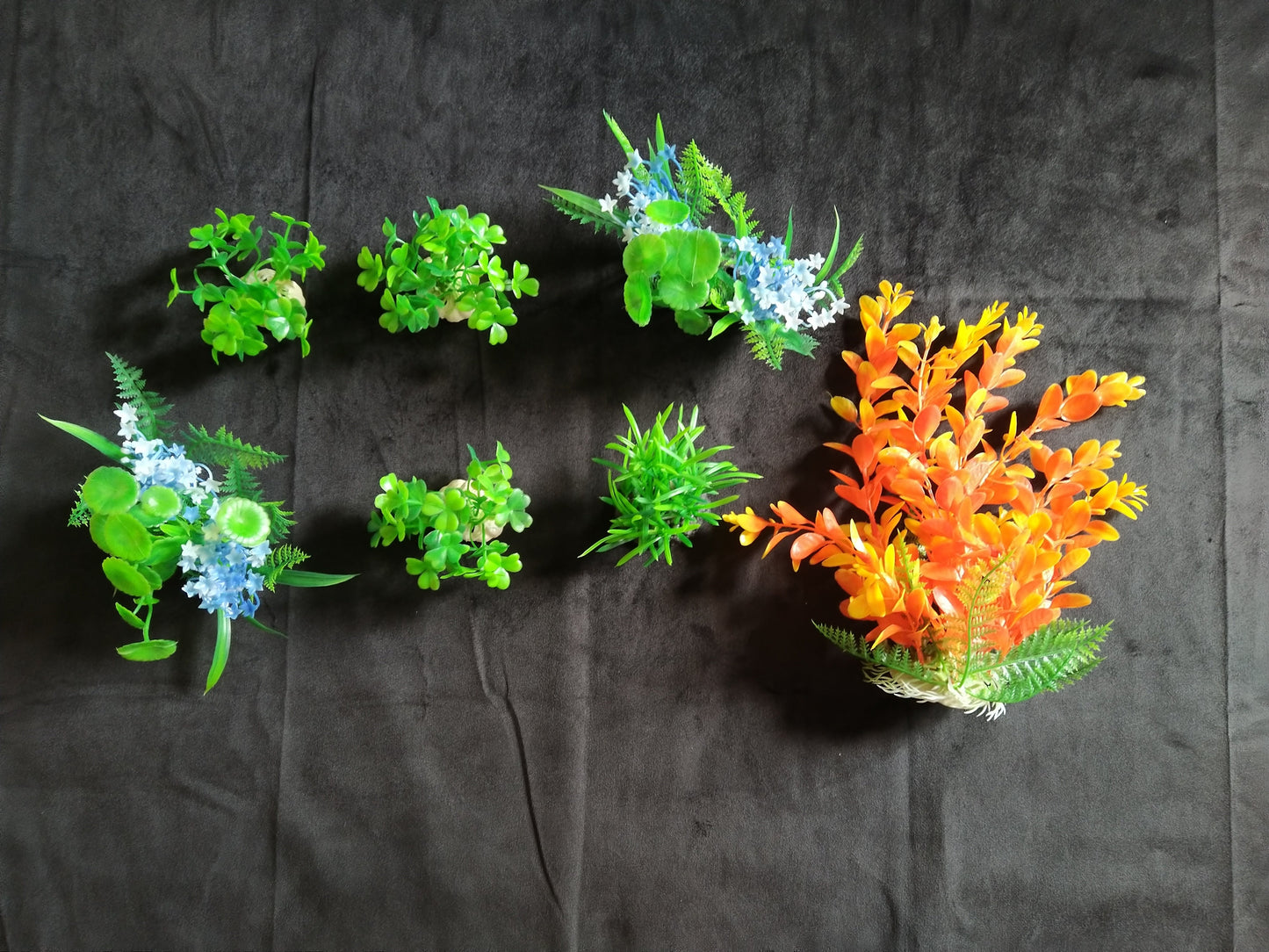 Set of artificial plants and flowers arranged in a circular pattern on a dark fabric background.
