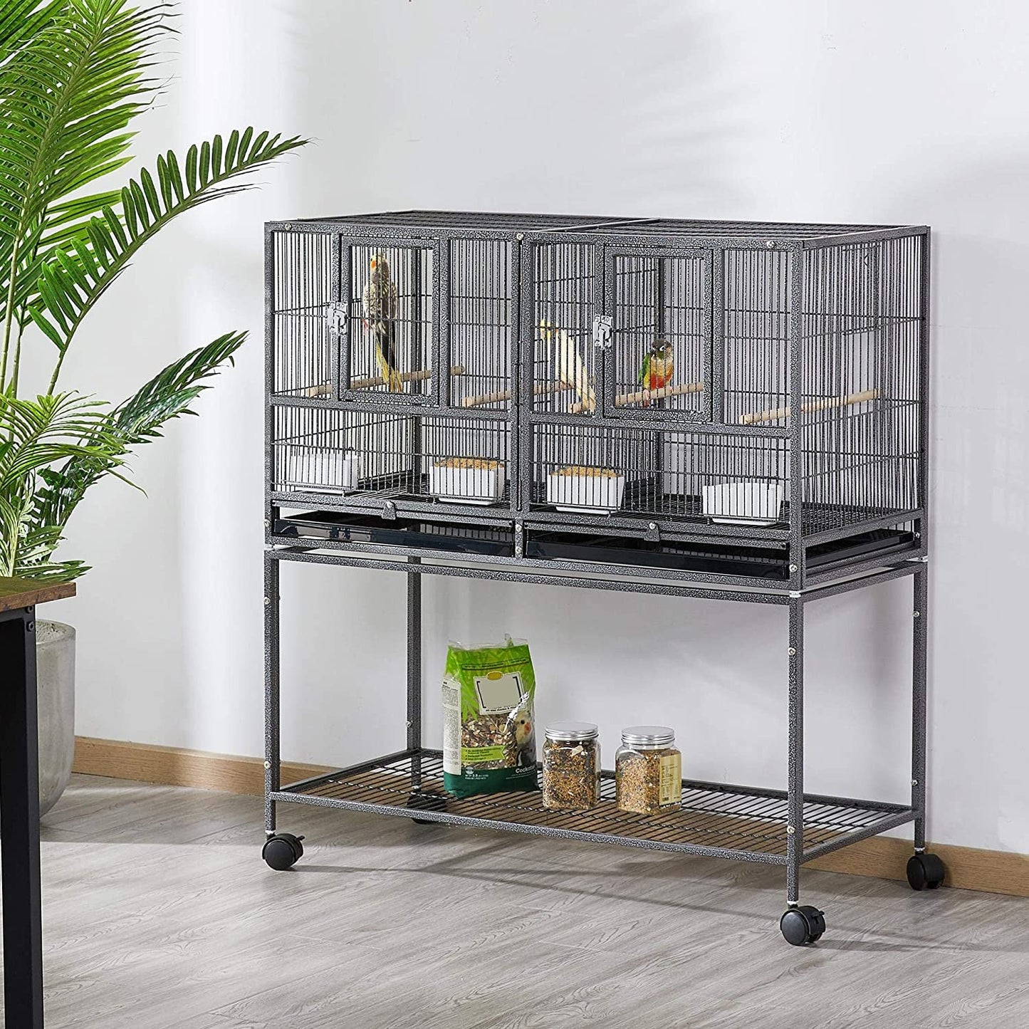 Large bird cage with multiple compartments on a stand, featuring wheels, in a room with a plant and pet food.