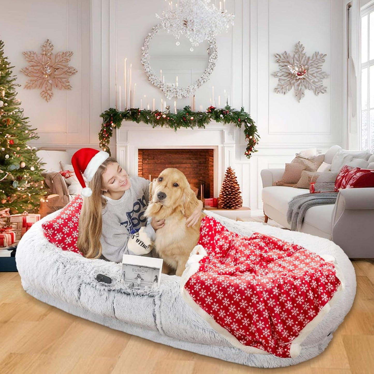 Woman in a Santa hat with a dog on a large pet bed in a festively decorated living room.