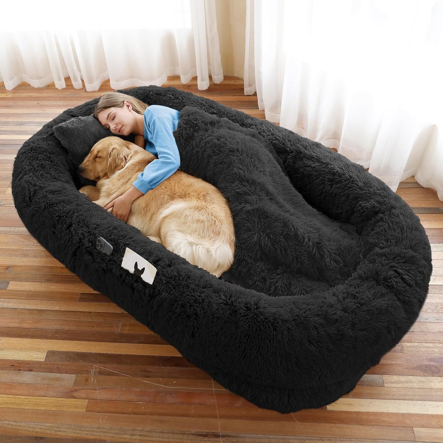 Woman and dog lying in a large black fluffy pet bed on a wooden floor.