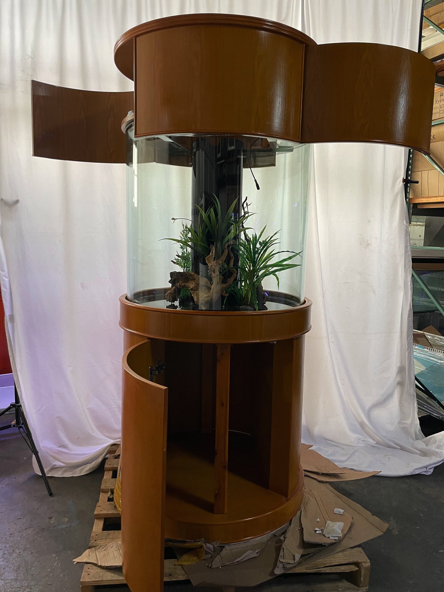 Brown cylindrical plant stand with plants inside, on a pallet against a white curtain background.