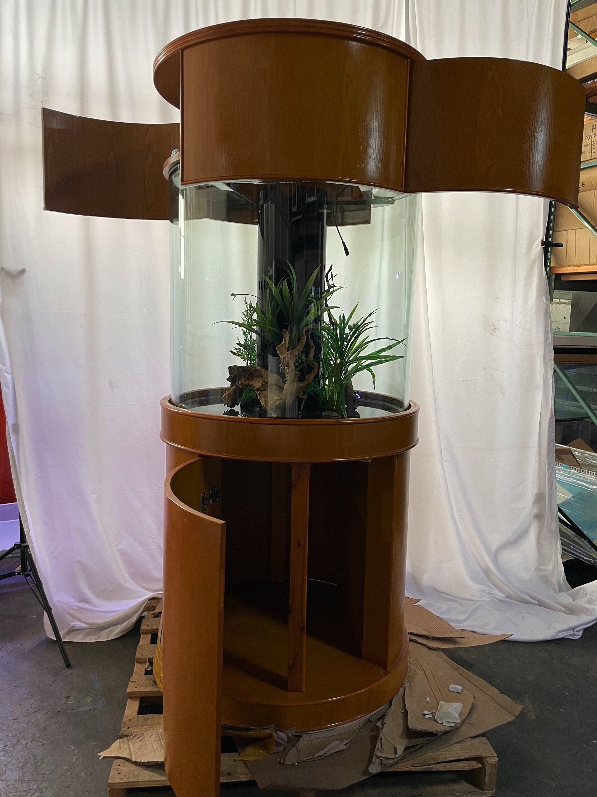 Brown cylindrical plant stand with plants inside, on a pallet against a white curtain background.