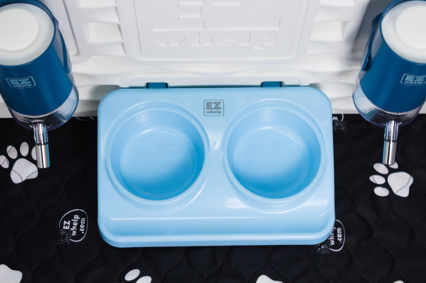Blue pet food and water bowls with a container on a black mat with paw prints.