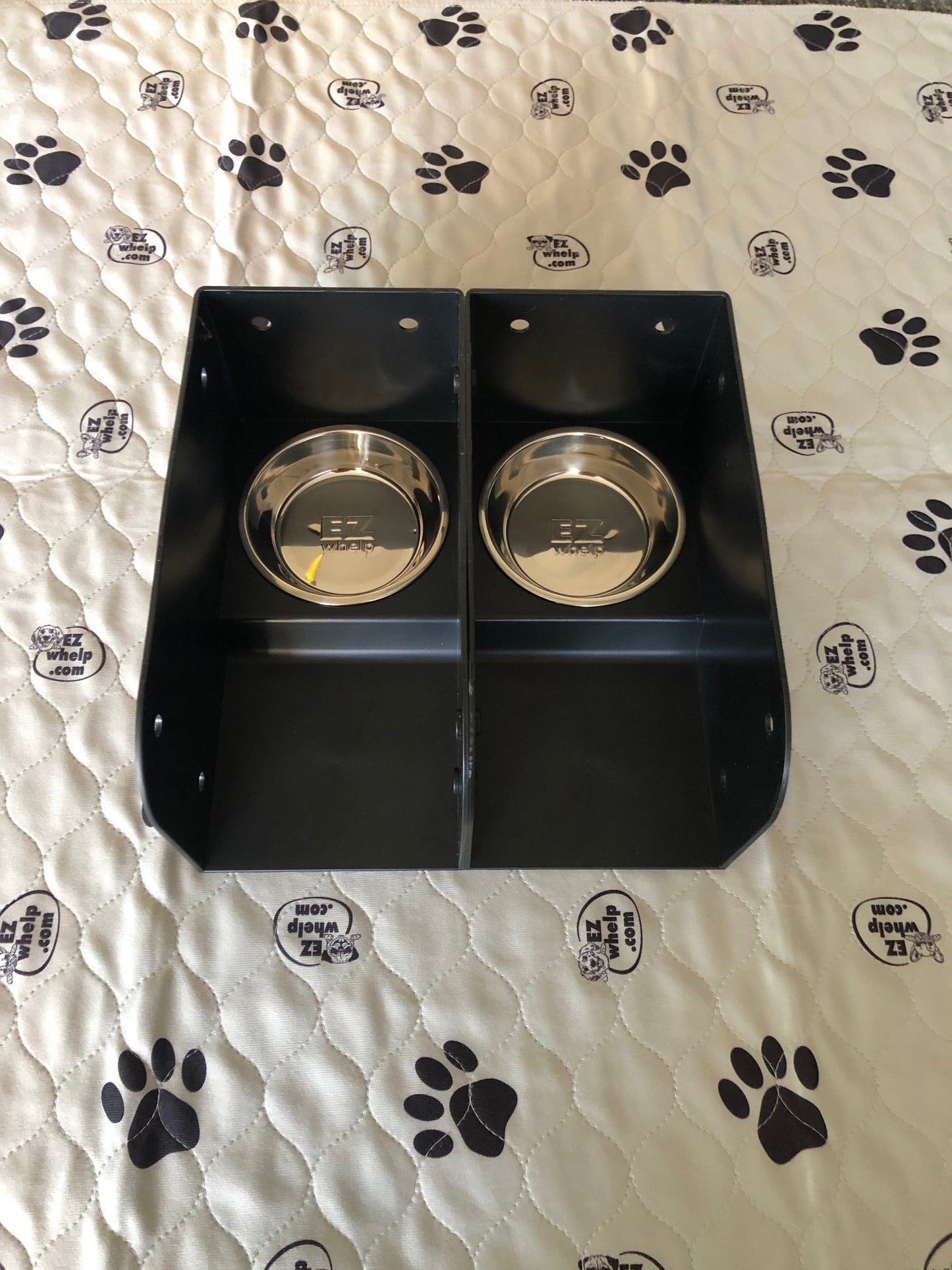 Black pet feeding station with two silver bowls on a white blanket with black paw prints.