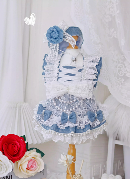 Doll in a blue and white dress with floral accessories against a white curtain background.