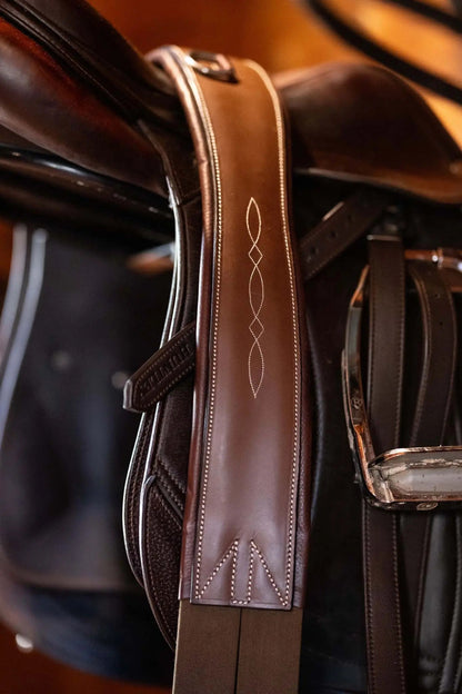 Close-up of a brown leather saddle with intricate stitching on a wooden floor.
