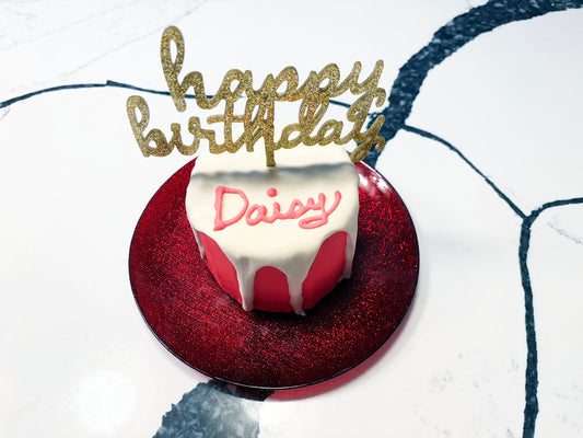 Small round cake with white icing and red base, topped with a 'Happy Birthday Daisy' gold topper.