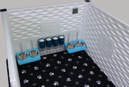 Pet crate with bedding, water bottles, and bowls on a black paw print mat.