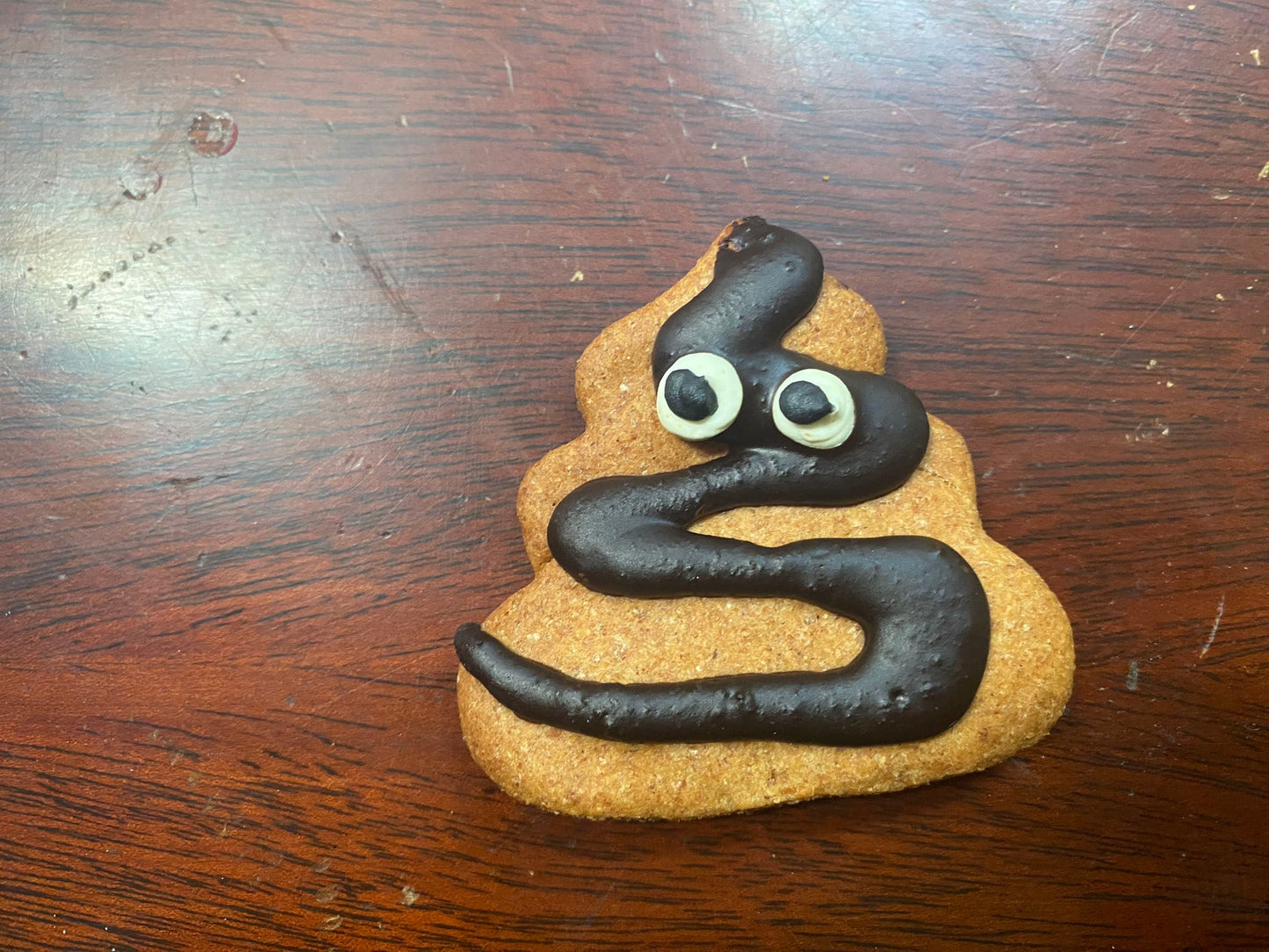 Cookie shaped like a poop emoji with chocolate 'poop' and candy eyes on a wooden surface