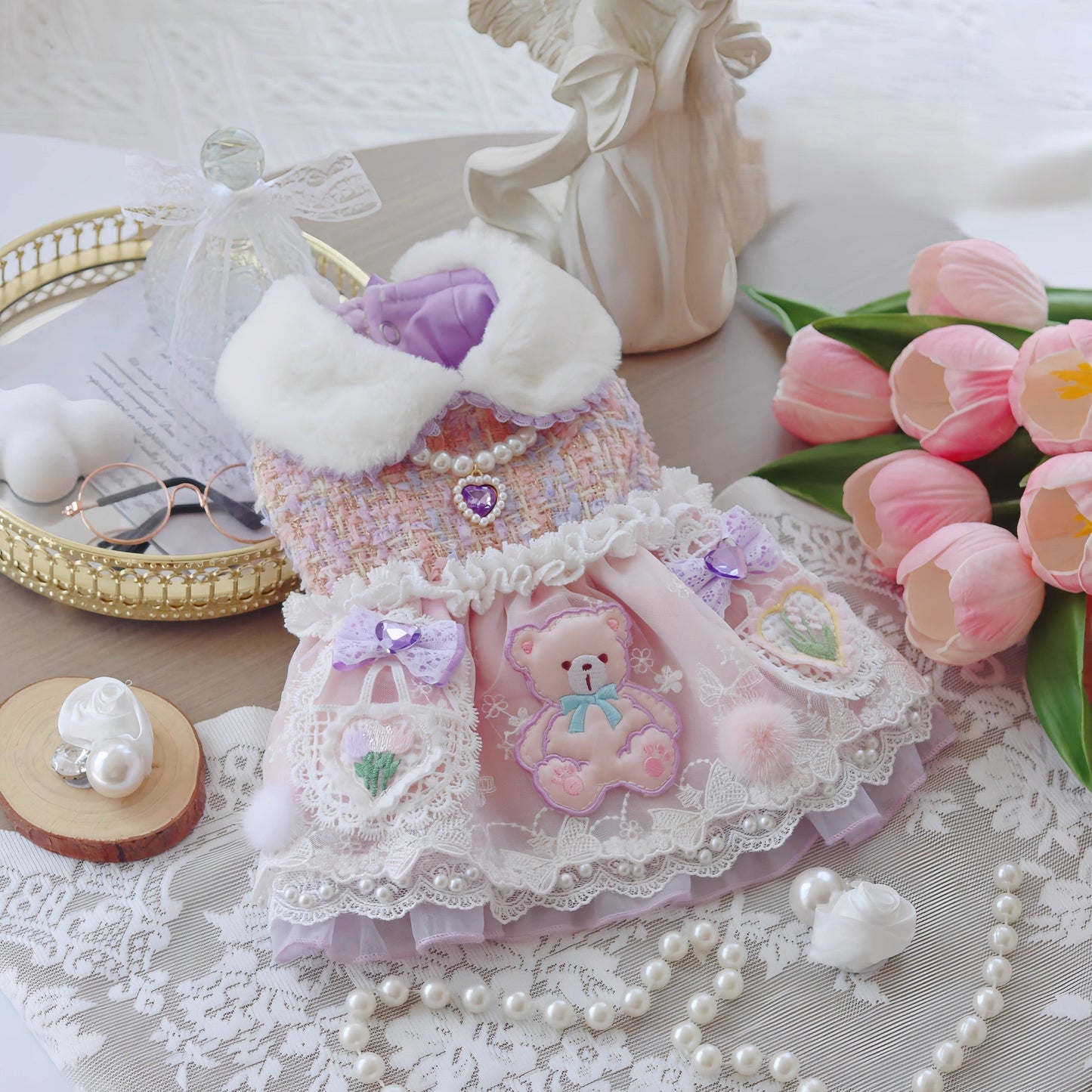 Delicate pink dress with teddy bear design on a lace tablecloth with flowers and jewelry.
