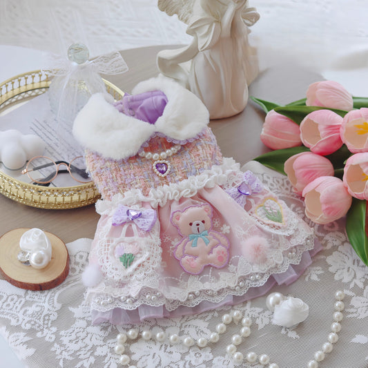 Delicate pink dress with teddy bear design on a lace tablecloth with flowers and jewelry.