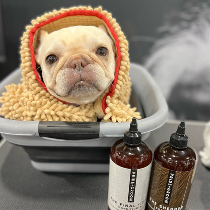 Dog in a bath with a towel on its head, surrounded by pet grooming products.