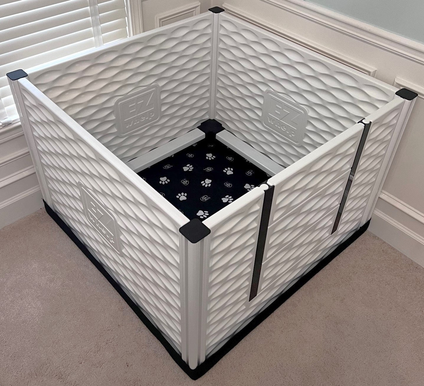 White pet playpen with a black mat inside on a carpeted floor.