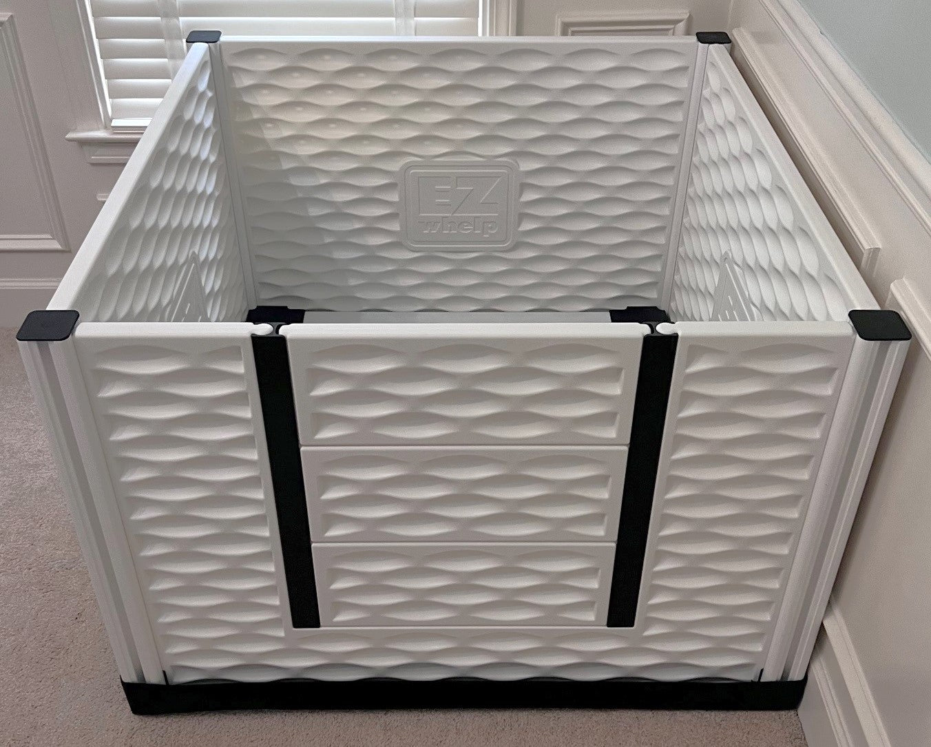 White pet playpen with black straps in a room with light-colored walls and carpet.