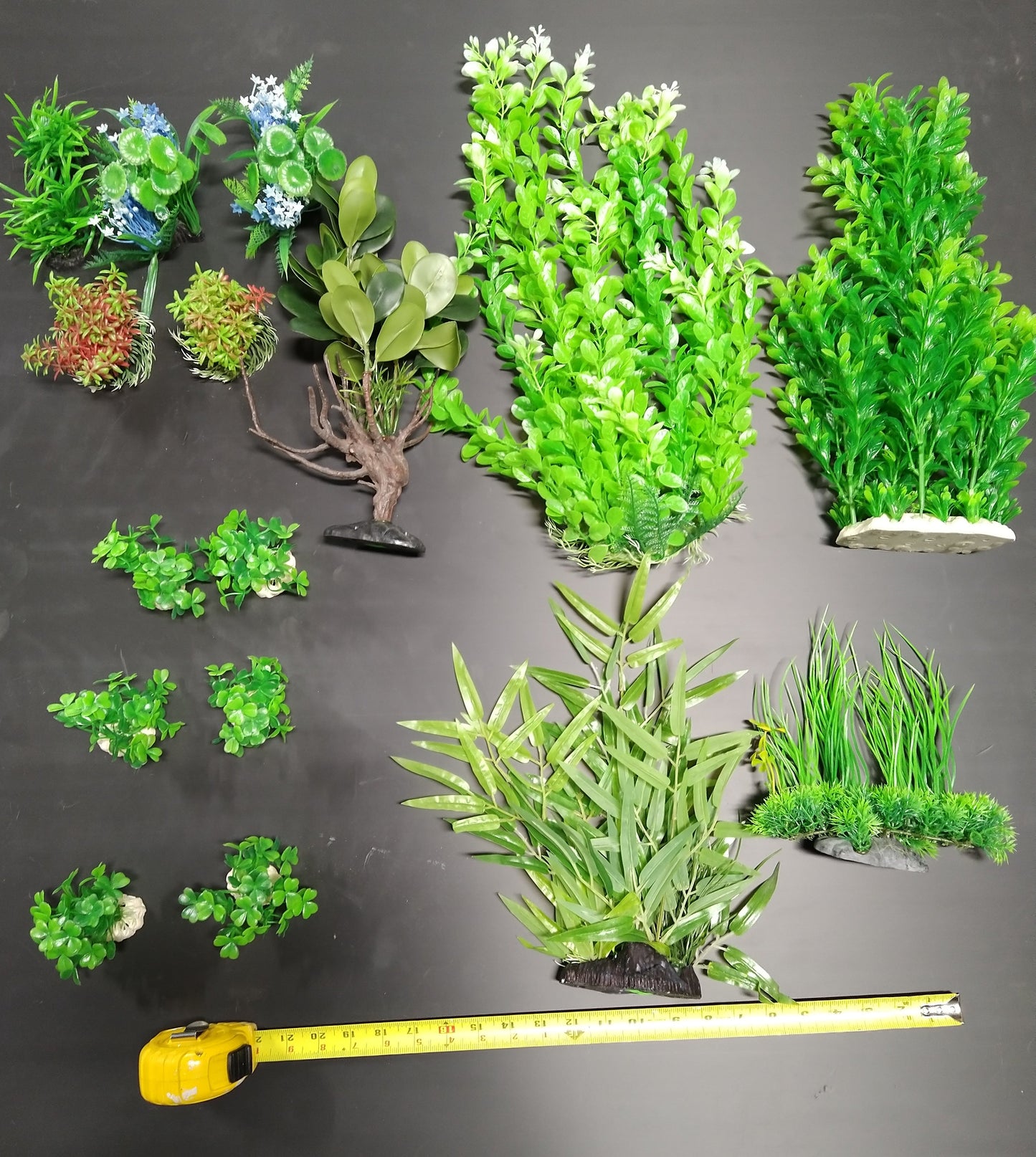 Assorted artificial green plants of various sizes on a dark surface with a measuring tape for scale.