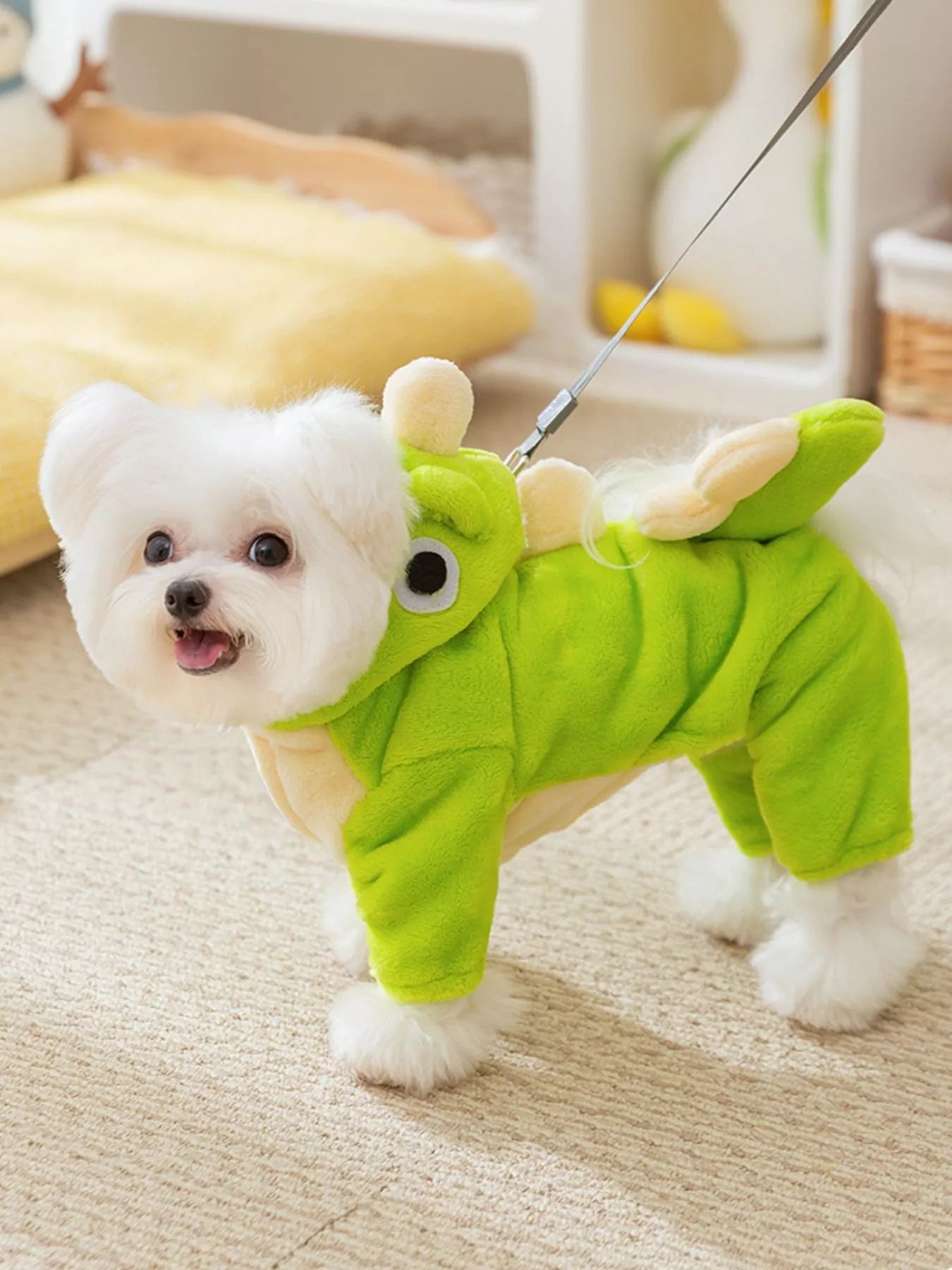 Small white dog wearing a green dinosaur costume indoors.