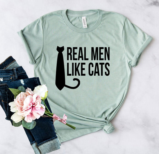 Mint green t-shirt with 'Real Men Like Cats' text and a cat tail graphic on a light gray background.