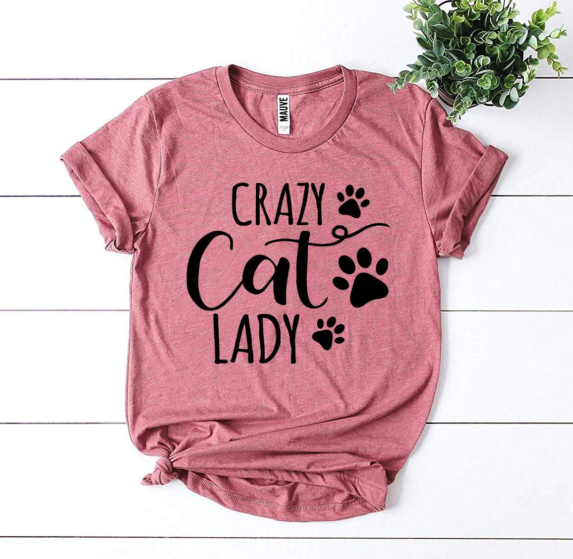 Pink t-shirt with 'Crazy Cat Lady' text and paw prints on a white wooden surface.