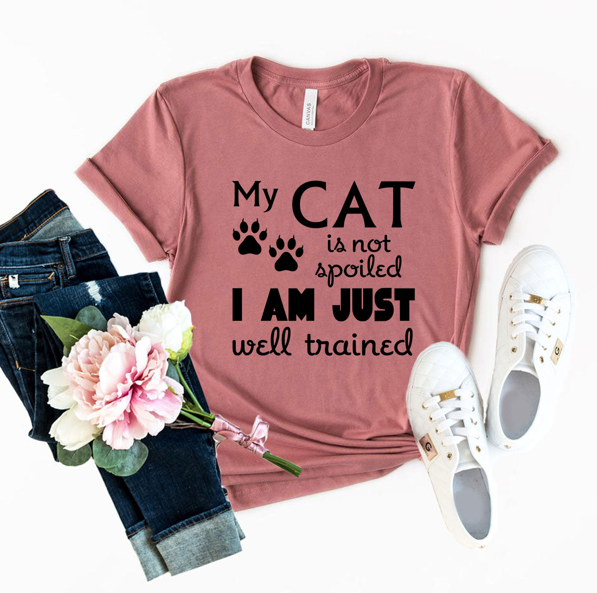 Pink t-shirt with 'My cat is not spoiled, I am just well trained' text, paired with jeans, sneakers, and flowers on a white background.