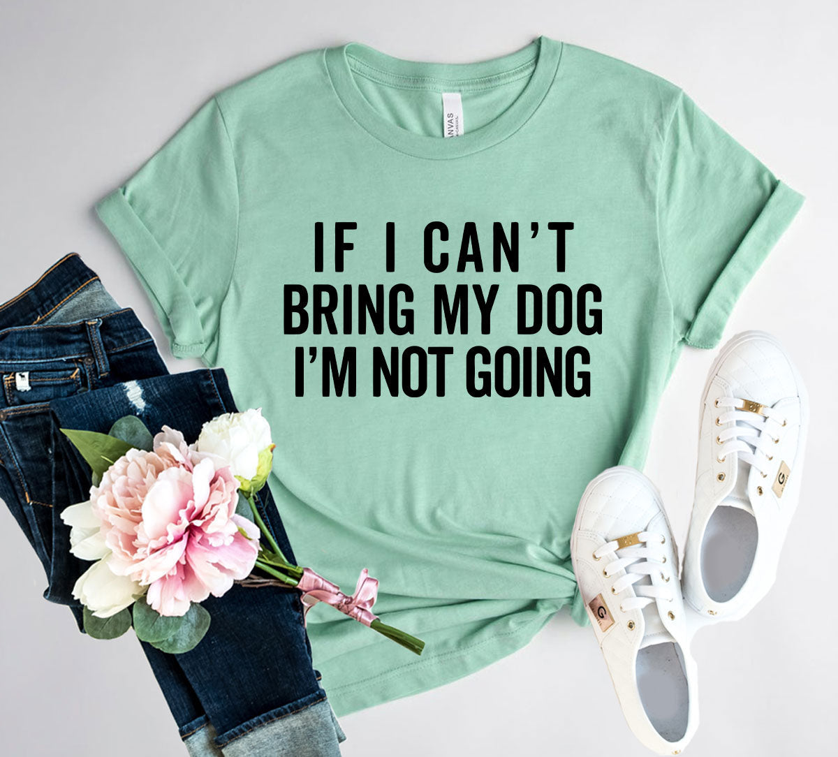 Mint green t-shirt with text 'If I can't bring my dog, I'm not going' on a light gray background with jeans, flowers, and sneakers.