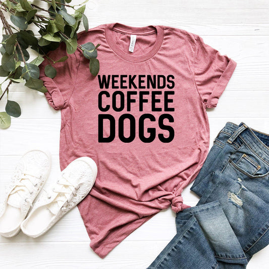 Pink t-shirt with 'Weekends Coffee Dogs' text on a white surface with jeans and sneakers.