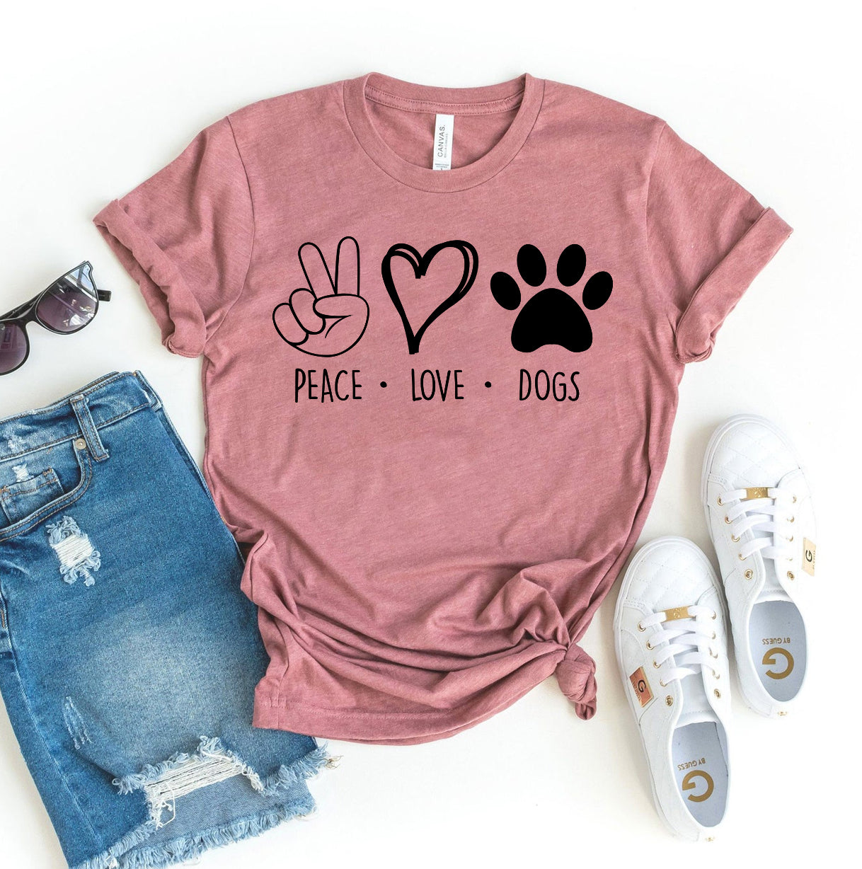 Pink t-shirt with 'Peace Love Dogs' design, sunglasses, denim shorts, and white sneakers on a white background