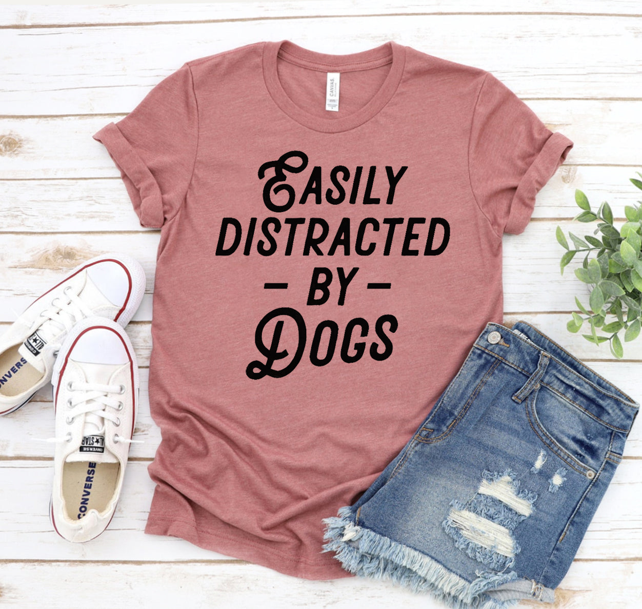 Mauve t-shirt with 'Easily Distracted by Dogs' text, paired with denim shorts and white sneakers on a wooden surface.