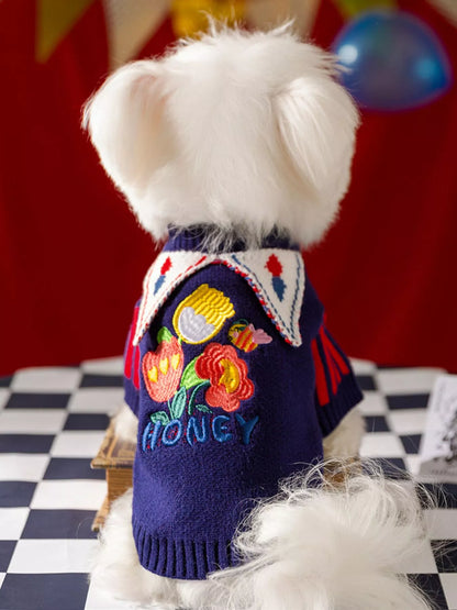 White dog wearing a colorful sweater with floral designs and the word 'Honey' on a checkered floor.