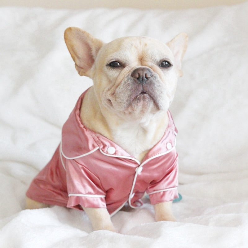 Dog wearing a pink satin pajama outfit on a white bed.