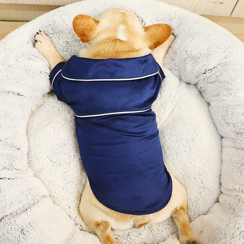 Dog wearing a blue pajama outfit lying on a fluffy white bed.